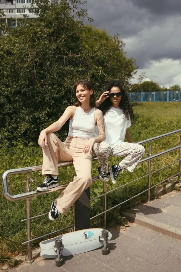 Duas amigas sorrindo sentadas em corrimão de parque com skate, uma delas de camiseta branca lisa