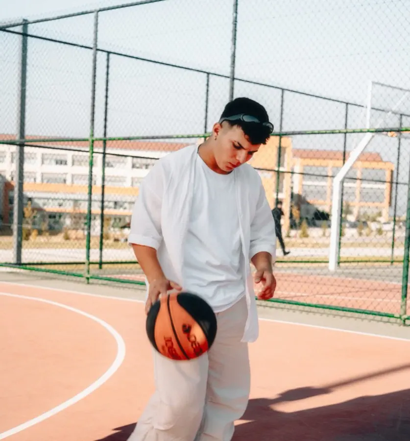Jovem de camiseta oversized branca driblando bola de basquete em quadra externa laranja