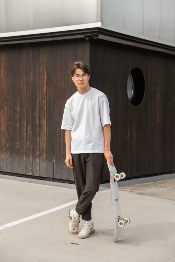 Jovem de camiseta oversized branca lisa segurando skate em frente a fachada de madeira moderna