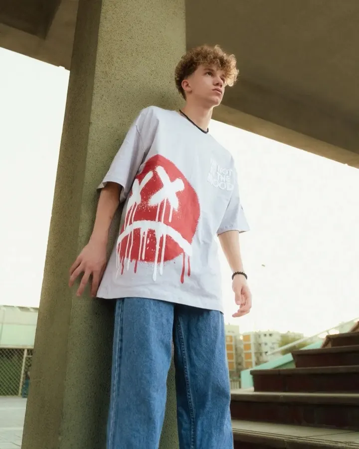 Jovem vestindo camiseta oversized cinza com estampa moderna de smiley vermelho, encostado em pilar de concreto