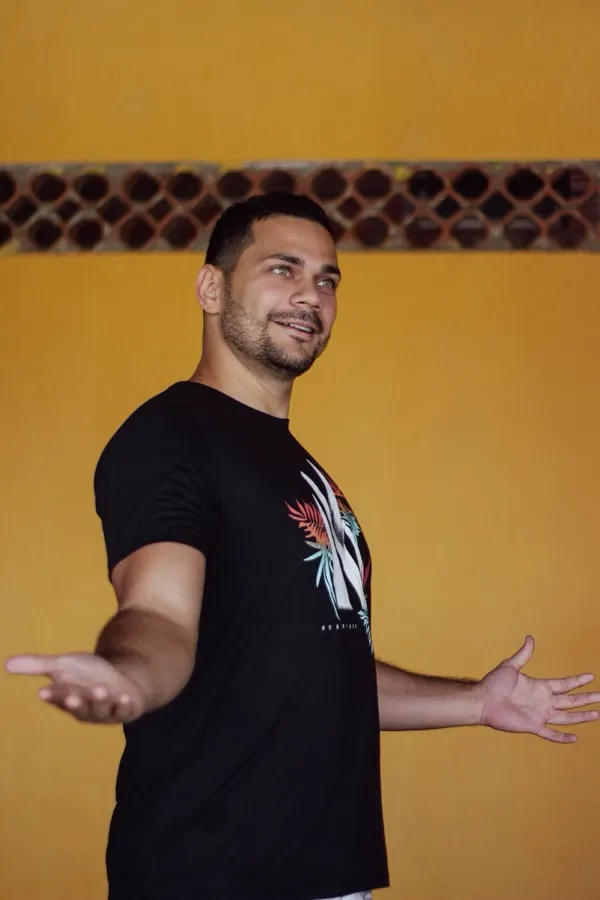 Homem sorrindo de braços abertos com camiseta preta de estampa tropical em frente a parede amarela vibrante